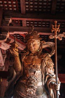 Tahta bir muhafız, Nara Tapınağı 'nın içinde bir mızrak değneği ve küçük bir pagoda tutuyor. Ilık ışıkla aydınlatılan süslü zırh ve taç. Ölçek ve doku için yakın ve yukarı bakıyor..