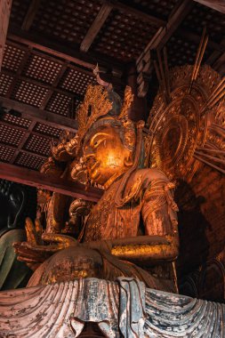 Altın yaldızlı bir Budist heykeli Japonya 'nın Nara şehrinde iç ışık altında parıldıyor. Süslü taç, haleler, katmanlı bornozlar ve el mudroları yakından yukarı doğru görünüyor..