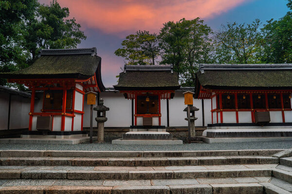 Три маленьких павильона Синто с вермилионными колоннами стоят во внутреннем дворике Киотского храма в сумерках. Каменные фонари, гипсовые стены и гранитные лестницы обрамляют спокойную церемониальную сцену.