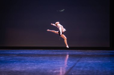 Profesyonel bir bayan dansçı loş bir sahnede dramatik bir jete atlar. Tek bir spot ışığı, parlak zeminde yansımalar göründüğünde kemerli şeklini vurgular..