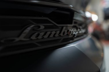 Dar bir alçak açı, parlak siyah bir Lamborghini arka rozeti keskin odak, bulanık atölye araçları ve arkada asansörler, karamsar ışıklandırma ve sığ alan derinliği gösterir.