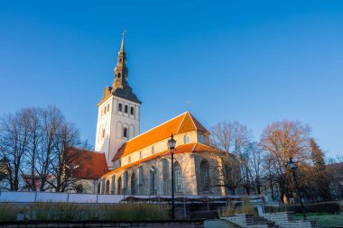 Tallinn Eski Kasabası 'ndaki Aziz Niklas Kilisesi yapraksız ağaçların ve taş bir duvarın üzerinde yükselir. Turuncu kiremitler, beyaz bir kule, Gotik detaylar ve uzun gölgeler kışın keskin manzarasını tanımlar..