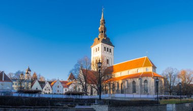 Güneşli Aziz Niklas Kilisesi kışın Tallinn Eski Kasabası üzerinde yükselir. Uzun bir kule, turuncu çatı, Gotik naf, çıplak ağaçlar, sokak lambaları ve soğan kubbeleri soğuk bir festival sahnesini tanımlar..