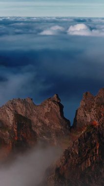 Sivri uçlu Pico do Arieiro tepeleri Portekiz 'in Madeira Adası' ndaki yoğun bulut dönüşümünü deliyor. Sis, şafak veya alacakaranlıkta kayalıklarda süzülürken, alçak sıcak ışık kırmızı kehribar kayalıklarını boyar..