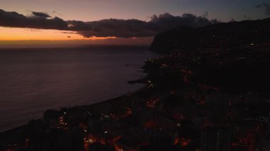 Funchal, Madeira, Portekiz 'in hava alacakaranlık manzarası, sağda bir uçurum, solda sakin yansıtıcı Atlantik, turuncu günbatımı bandı, sokak ışıkları, pencereler ve küçük bir iskele..