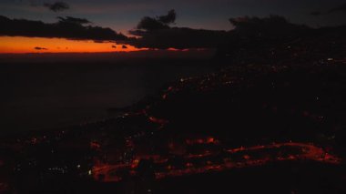 Portekiz, Madeira Adası, Funchal 'ın hava alacakaranlık manzarası. İnsansız hava aracı caddeleri süpürürken virajlı yolları izler, uçurumlar sağdan yükselir, sakin deniz solda uzanır, ufuk turuncu parlar..