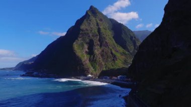 Hava görüntüsü Madeira 'nın kuzey kıyısı bazalt kayalıkları, siyah kum körfezi, sahil köyü, beyaz pavyon, yol ve rıhtım, turkuaz sörf ve hafif hareketli güneşli kayalıklar gösteriyor..