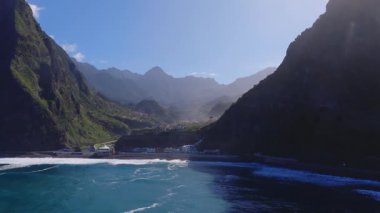 Hava görüntüsü, Madeira, Portekiz 'de yeşil uçurumları, teraslı evleri, deniz manzaralı bir köşkü, alçak bir köprüsü, siyah kumu ve gün ışığında dalgaları olan bir kıyı köyünü gösteriyor..