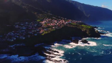 Hava manzarası Portekiz 'in Madeira adasında kıyı köyü gösteriyor. Teraslı kayalıklarda kırmızı fayanslı evler, volkanik kayalar, turkuaz Atlantik sörfü ve öğleden sonra ışığı var.