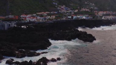 Hava kapları Porto Moniz lav havuzları, bazalt kayalıkları ve Madeira 'da deniz kenarı gezinti alanı gösteriyor. Dalgalar çarpar, sörfler havuzları doldurur, ziyaretçiler güneşlenir ve gece yarısı ılık ışıklarda dolaşır..