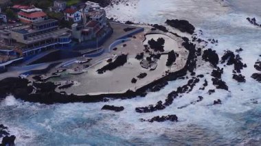 Hava aracı görüntüleri Madeira 'nın volkanik kıyısındaki Funchal Lido havuzlarını gösteriyor. Yürüme yolları, teraslar, dalgalar çöktüğünde binalar ve insanlar öğleden sonra ışıklarında geziniyor..