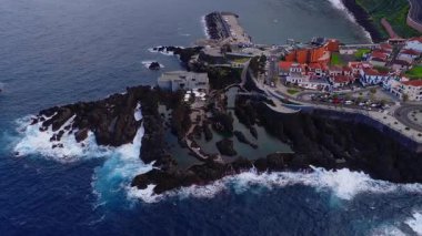 Hava görüntülerine göre Porto Moniz gelgit havuzları, dalgalar çarptığında sivri bazalt, liman ve gezinti alanları, tekneler hareket ediyor ve ziyaretçiler Madeira 'daki kırmızı çatı evlerinin yanından geçiyorlar..