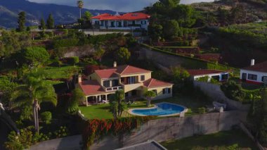 Havalandırma panosu Funchal, Madeira yakınlarında teraslı villalar gösteriyor. Sarı iki katlı villa, kırmızı kiremitli çatılar, bahçeler, böbrek havuzu, palmiye ağaçları, sıcak ışık, uzun gölgeler..