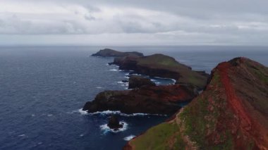 Hava kamerası Ponta de Sao Lourenco, Madeira, Portekiz boyunca süzülür. Pürüzlü kırmızı kahverengi kayalıklar, deniz yığınları, dar bir patikadaki yürüyüşçüler, dalgalar ve bulutlu ışık mevcuttur.