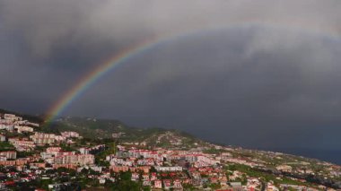 Funchal, Madeira, Portekiz 'in havadan görünüşü teraslı kırmızı çatılar, dik kıyı arazisi ve bulutlar kayarken canlı bir gökkuşağı ve güneş ışığı çatıları vurguluyor..