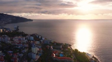 Funchal, Madeira 'nın havadan görünüşü rıhtıma bakan oteller, havuzlar, yolcu gemisi terminali, teraslı daireler ve katmanlı bulutlarla dolu sıcak altın saat arka planında engebeli uçurumlar gösteriyor..