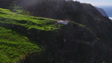 Hava görüntüsü Portekiz, Madeira 'da yeşil bir platoda çitli bir miradouro gösteriyor. Dalgalar öğleden sonra aydınlık ve puslu bir havada kayalıklara çarptığında insanlar el sallıyor..
