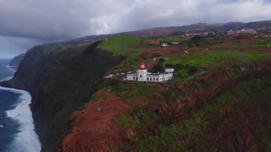 Portekiz, Madeira 'daki Farol da Ponta do Pargo' nun hava manzarası. İnsanlar beyaz deniz fenerinin ve uçurumun kenarında yürür dalgalar kopar ve bir dron pelerini süpürür.