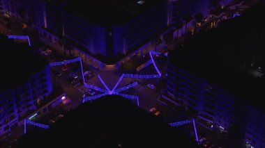 Funchal, Madeira 'daki hava gece manzarasında oteller ve daireler, mavi ve mor LED ışıklardan oluşan bir yıldız merkezi, hareket eden arabalar, yaya geçitleri, palmiye ağaçları ve bir çatı havuzu görülüyor..