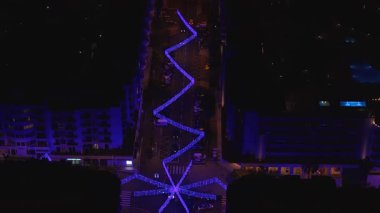 Hava görüntüsü Portekiz, Funchal 'da uzun bir bulvar gösteriyor. Mavi mor LED kurdeleleri bir kavşakta yıldıza yaklaşıyor. Oteller ve havuzlar gece aydınlanıyor..