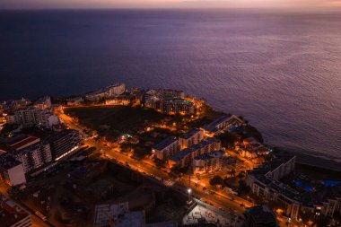 Madeira 'nın Funchal kıyı bölgesinin hava alacakaranlık manzarası, kavisli yollarda turuncu sokak lambaları, parkın yanındaki yükselen oteller, kayalık gezinti yerleri ve derin mavi Atlantik.