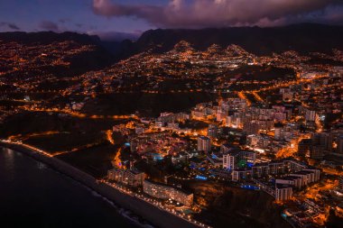 Funchal, Madeira, Portekiz 'in havadan gece görüşü, sokak lambalarıyla aydınlatılmış teraslı tepeler ve vadiler, dik yamaçlardaki dolambaçlı yollar, zirvelerdeki bulutlar ve dar bir gezinti alanı..