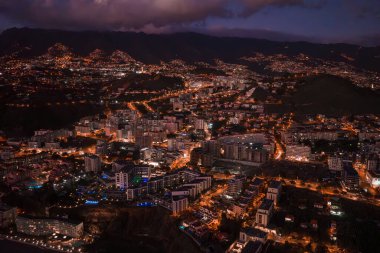 Aerial twilight view of Funchal, Madeira shows mid rise buildings, terraced hills, winding lit roads, dark mountains, low clouds, and evening activity with blue lit accents.