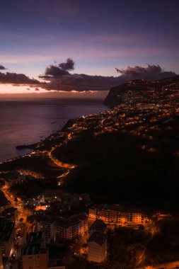Funchal yakınlarındaki Madeira güney kıyısındaki hava alacakaranlığı, sokak lambaları dolambaçlı yollar, teraslı kasabalar küçük limanlara iniyor, alçak bulutlar gün batımından sonra asılıyor..