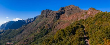 Portekiz 'in Madeira Adası' ndaki Pico do Arieiro 'da volkanik kayalıklar defne ormanlarının üzerinde yükselir. Yüksek kontrastlı gün ortası ışığı, Pico Ruivo 'ya doğru giden yolları ve katmanları ortaya çıkarır..
