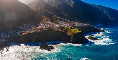 Hava görüntüsü Portekiz 'in Madeira şehrinde, yüksek kontrastlı ışık altında siyah bazalt' a çarpan turkuaz dalgalar nedeniyle teraslı tarlaları, beyaz evleri ve kırmızı çatıları olan bir uçurum köyü gösteriyor..