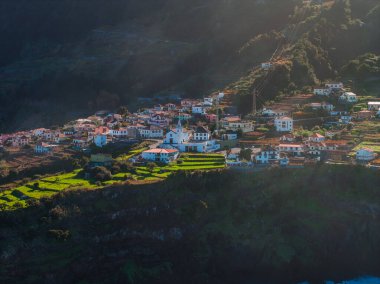 Beyazlatılmış evler Madeira, Portekiz 'deki bir kilisenin etrafında toplanıyor. Teraslar bir vadiye ve Atlantik kıyısına çıkar. Orman dağları ve öğleden sonra ışığı vardır..
