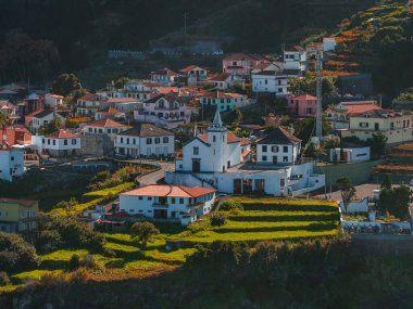 Portekiz 'in Madeira şehrinde bir çan kulesi olan bir kilisenin etrafında teraslı evler, öğleden sonra saatlerinde kıyı uçurumuna doğru ilerlerken, teraslı evler teraslı çan kulesi ile çevrili..