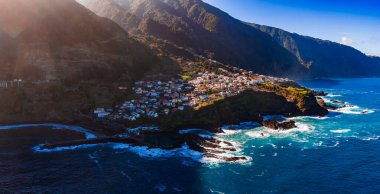 Hava görüntüsü Portekiz 'in Madeira Adası' nda bir uçurum sahili gösteriyor. Beyaz evler ve terracotta çatıları bir burunda, dağlar arkasında, sörf ve parlak öğleden sonra ışığı var..