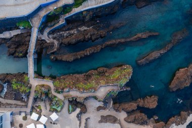 Yukarıdan bakıldığında Portekiz, Madeira 'daki Porto Moniz volkanik havuzları görülüyor. Siyah lav kayaları, turkuaz lagünler, yürüyüş yolları, teraslar, şemsiyeler ve gün ışığında yüzücüler..