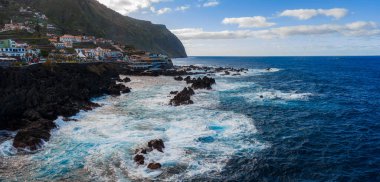 Porto Moniz, Madeira 'nın gündüz panoramik manzarası, siyah lav kayalarına çarpan dalgalar, yamaçtaki beyazlatılmış evler, yüksek bir burnu ve bulutların altındaki Atlantik suları..