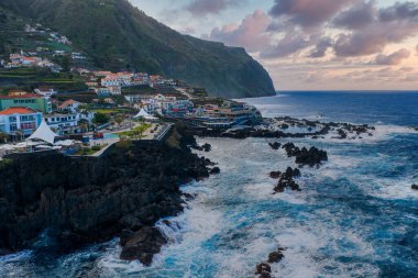 Porto Moniz, Madeira 'nın hava görüntüsü, Atlantik dalgalarının lav havuzlarına ve bazalt çıkıntılarına, dik kayalıklardaki teraslı evlere ve öğleden sonra geç saatlerde bir gezinti alanına vurduğunu gösteriyor..