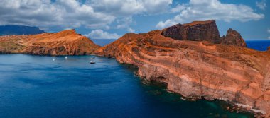 Hava görüntüsü, Portekiz, Madeira, Ponta de Sao Lourenco 'daki pas kırmızısı burunları gösteriyor. Volkanik kayalıklar, sakin bir körfez etrafında küçük tekneler, gün ışığı ve uzak tepelerle kıvrılır..