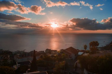 Sıcak gün batımı ışıkları Funchal, Madeira, teraslı evler, palmiyeler ve marina ipuçları, güneşi çerçeveleyen alçak bulutlar, ufukta kayıklar, geniş açı, geç gün ışığı.
