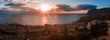 Funchal, Madeira Adası 'nın günbatımında geniş panoramik manzarası, güneş ışınları bulutları delip geçiyor, teraslı binalar limana iniyor, Cabo Girao sağa yükseliyor, tekneler yansıtıcı denizi gösteriyor..