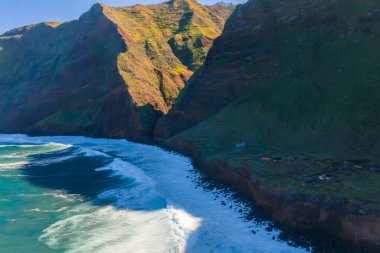 Gün ortası ışığı, Ponta de Sao Lourenco yakınlarındaki yeşil uçurumlara derin gölgeler düşürür. Engebeli kayalar, turkuaz sularla buluşur. Dar kayalıklar boyunca beyaz dalgalar kıvrılır..
