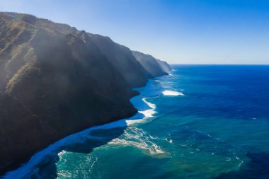 Hava görüntüsü Madeira 'nın kuzey kıyısı kayalıklarını, bazalt yamaçlarını ve kabarmalarını gösteriyor. Gün ortası ışığı, açık mavi gökyüzü ve Porto Moniz yakınlarında burunlar boyunca köpük çizgileri..