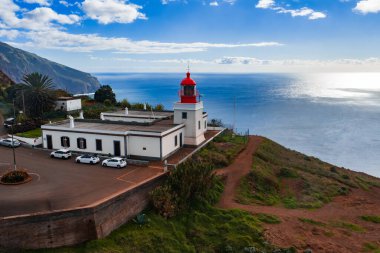 Ponta do Pargo, Madeira 'da kırmızı fenerli beyaz deniz feneri manzarası, küçük bir park yerinde arabalar, engebeli bir burunda kırmızı toprak yol, Atlantik üzerinde bulutlu gün ışığı..