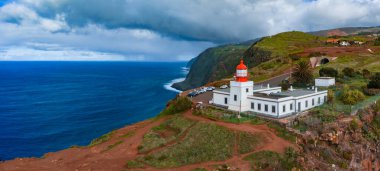 Hava görüntüsü Madeira, Portekiz 'deki yüksek uçurumlarda kırmızı kubbeli beyaz deniz feneri, dolambaçlı yol ve teraslı yamaçların yakınındaki park edilmiş arabalar, Atlantik üzerinde bulutlar dönüyor.