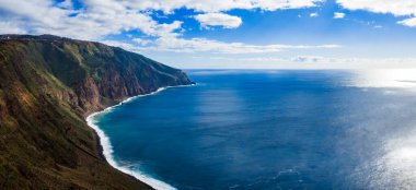 Beyaz dalgalar Ponta do Pargo, Madeira yakınlarında uzun bir burnun izini sürerken, yüksek uçurumlar mavi Atlantik ile buluşuyor. Güneş ışığı bulutları ve açık gün ortası kontrastı denizi ve kayayı tanımlar.