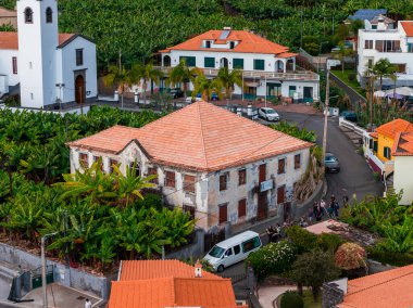 Hava görüntüsü, muz tarlaları, terracotta çatıları, çan kulesi, palmiye meydanı, park edilmiş arabalar ve Portekiz 'in Madeira kentinde minibüsü olan bir Madeiran köyünü gösteriyor..