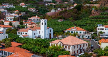 Portekiz, Madeira 'da kare bir çan kulesi olan, bembeyaz yıkanmış bir kilisenin üzerinde teraslı muz tarlaları yükseliyor. Kırmızı çatılar ve dar sokaklar ılık gün ışığında ve canlı renklerde görünür..