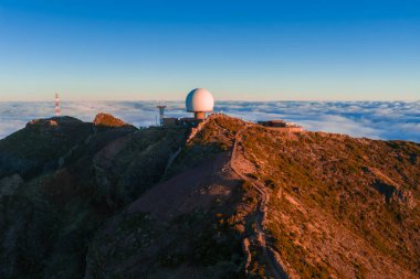 Hava görüntüsü Pico do Arieiro, Madeira, Portekiz 'de radar kubbesi, kuleler ve tepe izleri gösteriyor. Güneş doğarken bulutların üzerinde, volkanik yamaçlarda ve zirvede sıcak ışık var.