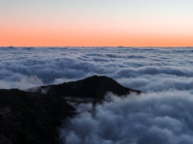 Portekiz 'in Madeira şehrinde güneş doğarken bulutların üzerinde bir dağ sırtı yükselir, Pico do Arieiro yakınlarında, yumuşak yayılmış bir ışık ve minimalist bir siluet manzarası vardır..