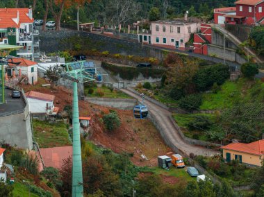 Funchal yakınlarındaki bir Madeira yamacının havadan görünüşü, dolambaçlı bir yolun üzerinde süzülen bir teleferik, teraslı bahçeler ve turuncu çatılar bulutlu bir ışık altında yeşil bir kulenin yanında oturuyor..