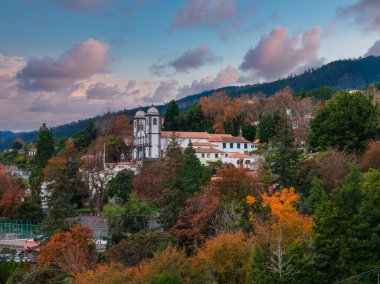 Monte, Madeira 'da kırmızı kiremitli çatıların üzerinden beyaz bir ikiz kilise yükselir. Gün sonunda bile serin, pembe bulutlar, sonbahar ağaçları ve katmanlı dağlar manzarayı oluşturur..